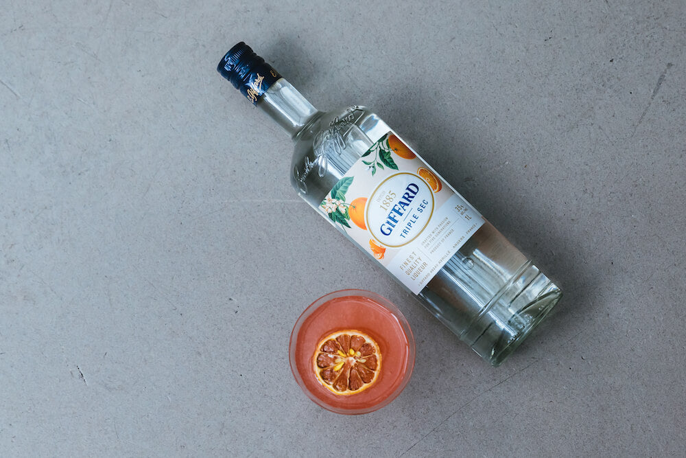 An overhead shot of a bottle of Giffard Triple Sec with a pink cocktail next to it. A candied orange slice floats on top of the cocktail.