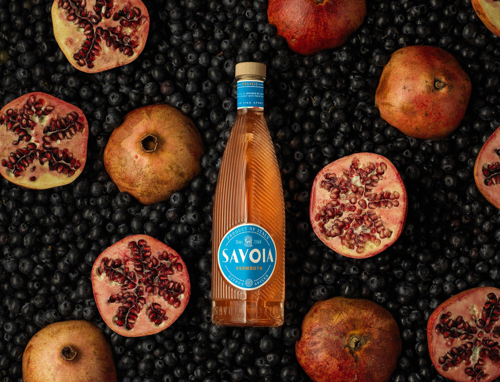 Bottle of Savoia Orancio laying on a bed of blue berries next to Pomegranates.