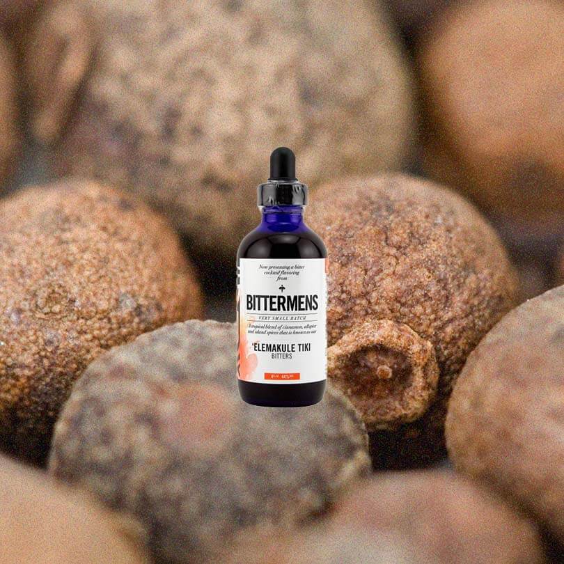 Bottle of Bittermens Elemakule Tiki Bitters against a macro shot of allspice.