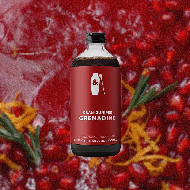 Bottle of Shaker & Spoon Cran-Juniper Grenadine against a delicious backdrop of a pomegranate and cranberry coulis, dotted by rosemary and orange peel.