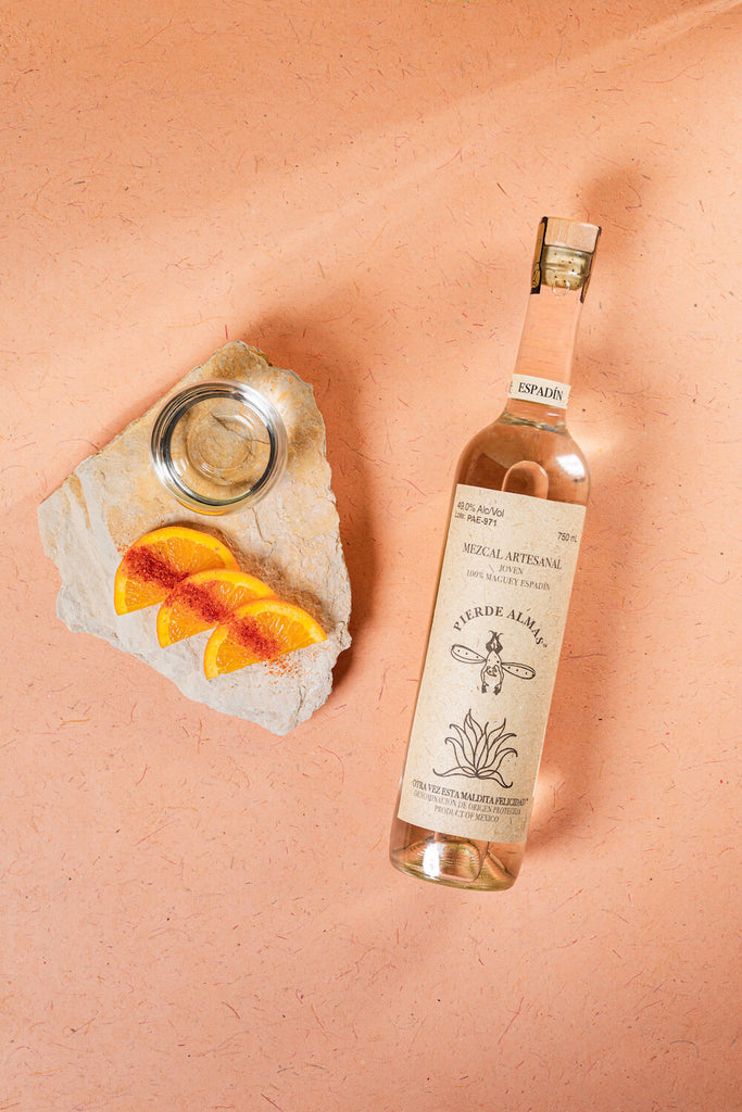 An overhead shot of Pierde Almas Mezcal next to a line of 3 orange slices dusted with taijin, all on a pleasing pink backdrop.