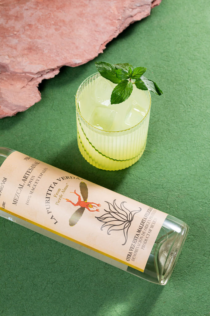 A close-up shot of the La Puritita Verda Mezcal Artesanal bottle on its side, a refreshing looking cocktail garnished with a mint leaf beside it. On a backdrop of green and pink.