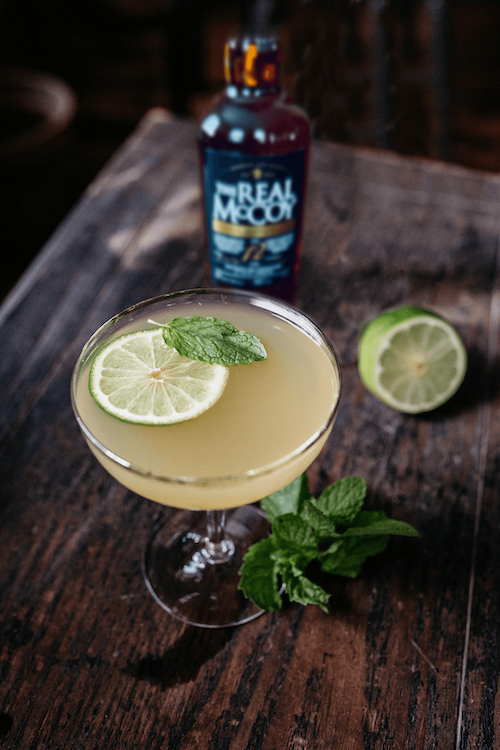 A beautiful pale yellow cocktail in a coupe with a lime wheel and mint floating on top. Likely a Daiquiri.  A Bottle fo The Real McCoy Single Blended 12 Year rum in the background.