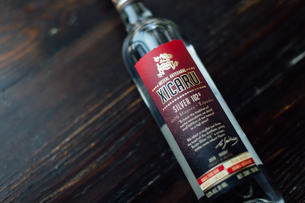 A hero image of Xicaru Silver 102, a close-up of the label reading Mezcal Artesanal. "To honor the tradition of master mezcaleros, we distille small batches of our mezcal to a high proof. We select a smaller cut from the heart of the distillate, giving Xicaru Silver 102 a crisper, richer, more robust flavor."