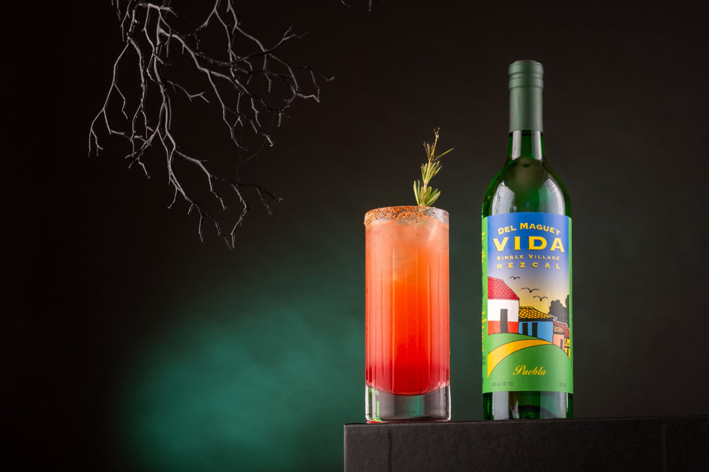 Bottle of Del Maguey VIDA Puebla Mezcal on a table next to a red cocktail with spooky Halloween themed background.