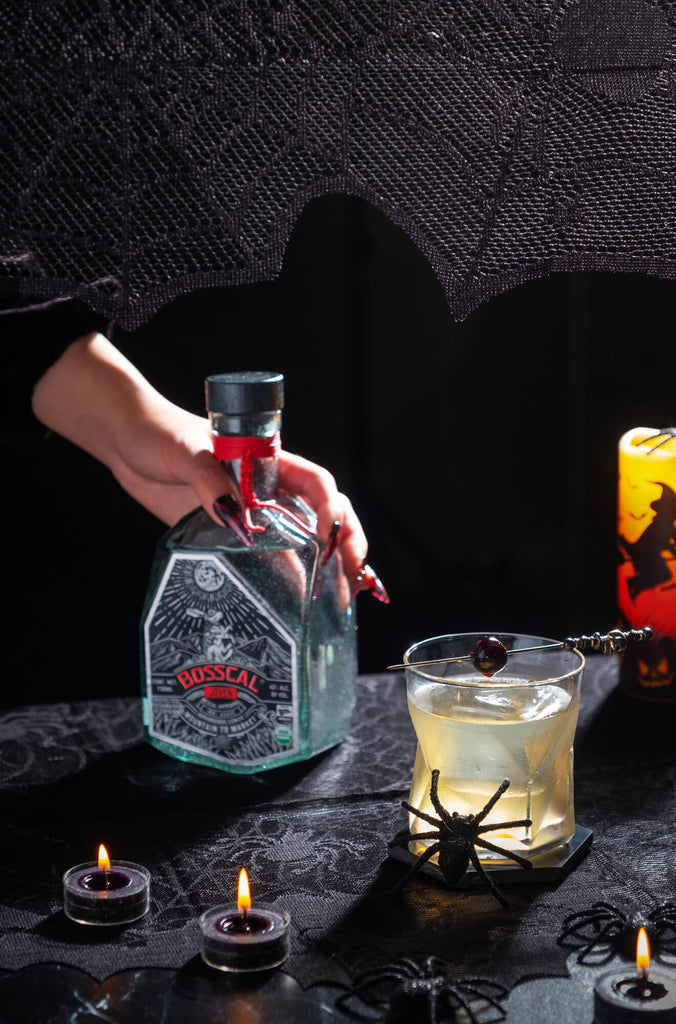 Person putting a bottle of Mezcal Bosscal Joven down on a table next to a cocktail. The table is decorated with Halloween decorations.