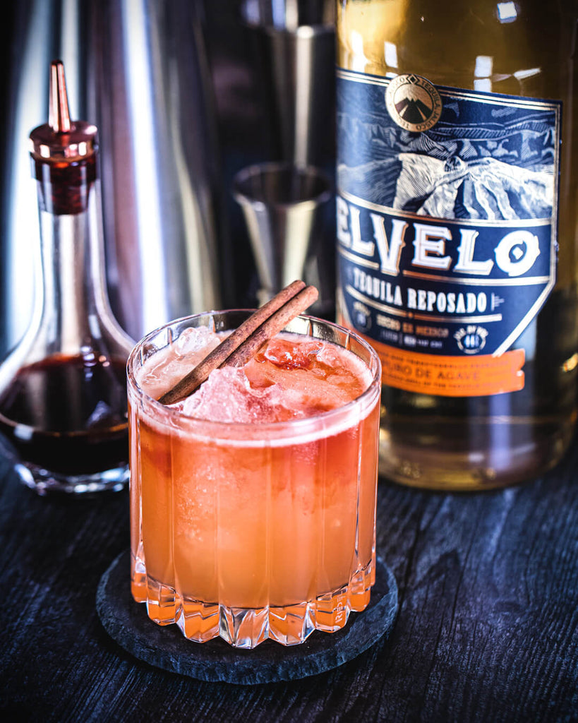 Light pink cocktail on dark blue table next to bitters and a bottle of tequila.