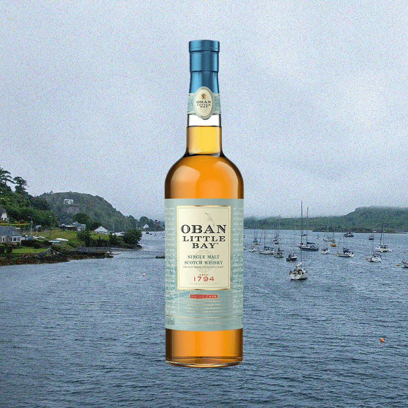 Bottle of Oban Single Malt Scotch Whisky Little Bay Small Cask over backdrop of sailboats anchored in the sea near the shore.