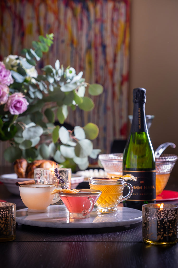 Bottle of Mumm Napa Brut Prestige on an elegantly set table with flowers, candles and three colorful cocktails garnished with spices.