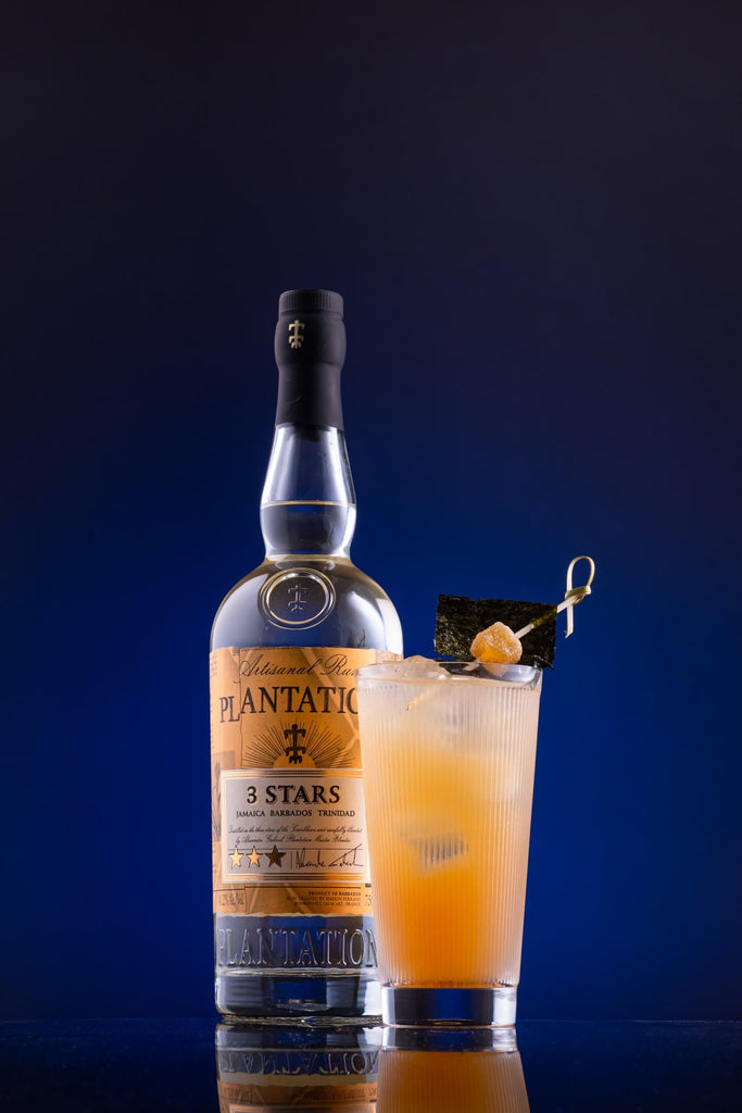 Bottle of Plantation 3 Stars Artisanal Rum with a navy blue backdrop and a light cocktail.