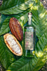 The angle is close-up bird's eye, or flat-lay.  There is a haled Cacao bean resting next to the bottle of Copalli Cacao Rum, all laid out on a bed of green cacao leaves. The bottle is tall and elegant, the rum is clear, with a label that reads Copalli Cacao Single Estate Organic. The label, rich browns and reds, depicts cacao leaves and beans in illustration.