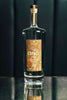 A tall elegant bottle of clear rum with a label that reads Copalli Cacao Single Estate Organic. The label, rich browns and reds, depicts cacao leaves and beans in illustration.  The bottle is set on a mirrored bar surface with a dark backdrop of a leather cafe booth, lending dramatic flair.