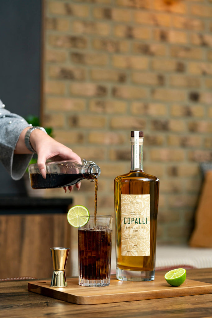 An image of a cocktail in a tall glass being prepared with a bottle of Copalli Barrel Rested rum to the glass's right. The cocktail is dark cola colored and has a lime garnish. It appears to be a kitchen backdrop with colors that are earthen, brick, and warm.