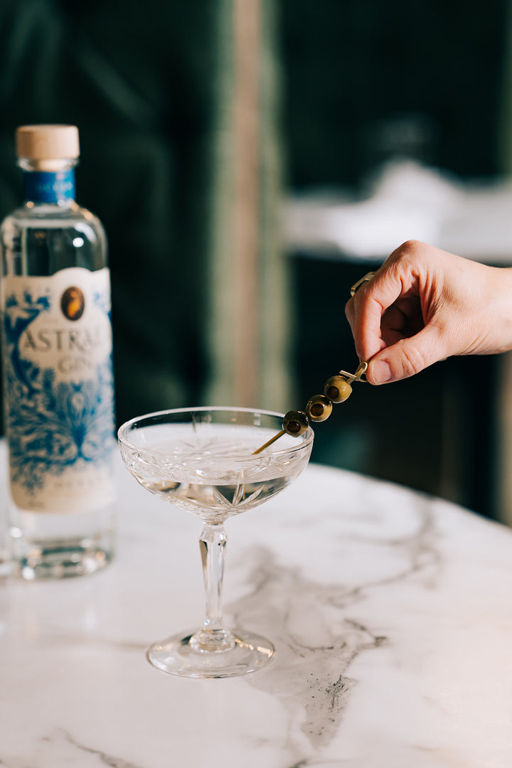 Person garnishing a cocktail with olives next to a bottle of Absolut Vodka on a marble surface.