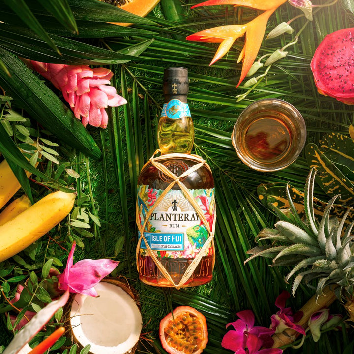 Bottle of Planteray Isle of Fiji Rum surrounded by tropical fruits and flowers on a green leafy background