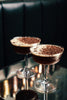 Two delicious looking cocktails in retro-looking coupe glasses, with dark liquid, a foamy top, garnished with coffee beans and cocoa powder. They are on a cafe table that is slightly mirrored, a dark banquette in the background.
