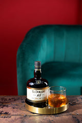 Bottle of El Dorado 15-Year-Old rum on a gold coaster with a glass of rum in front of a teal chair and red wall.