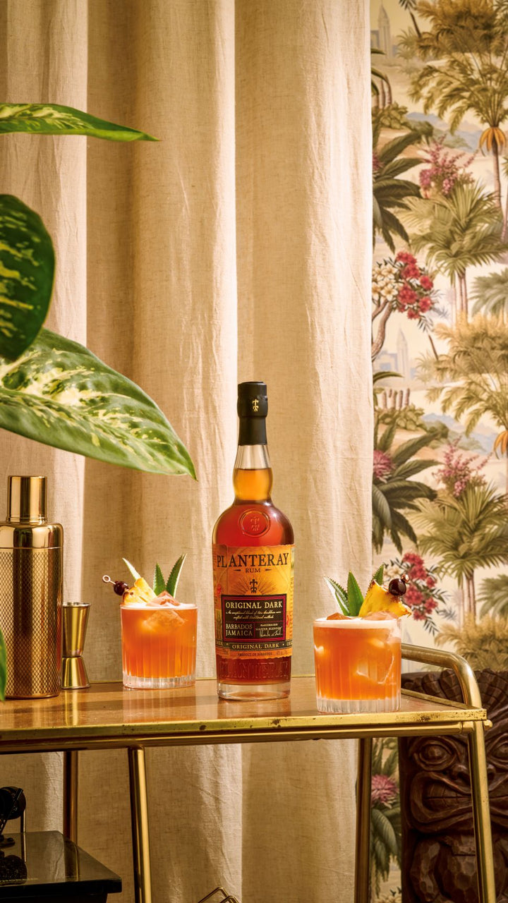Bottle of Planteray rum on a table with two cocktails and decorative elements.