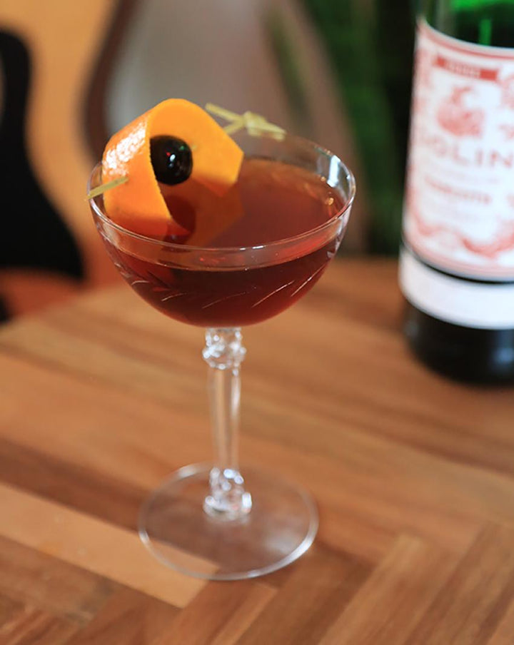 Cocktail in a glass with an orange twist on a wooden table, with a bottle in the background.