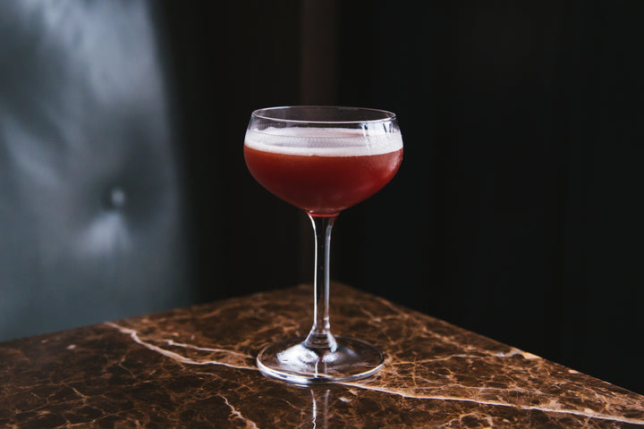 Red cocktail with a white foam top in a glass on a dark marble surface