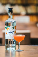A bottle of Giffard triple sec and a pink cocktail in sharp focus, a blurred setting in the background with warm colors.