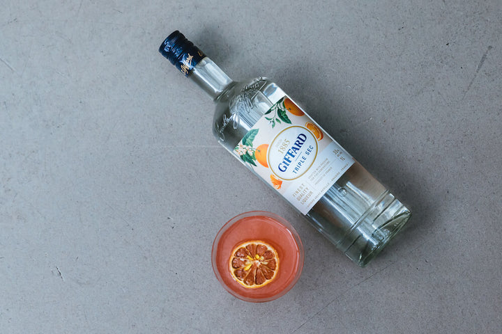 An overhead shot of a bottle of Giffard Triple Sec with a pink cocktail next to it. A candied orange slice floats on top of the cocktail.