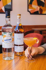 An interior shot of a hand holding a cocktail in a Nick and Nora glass. The person is seated a table in an artfully decorated restaurant. The foreground shows a bottle of Giffard Triple Sec. The cocktail is a pleasing hue of orange.