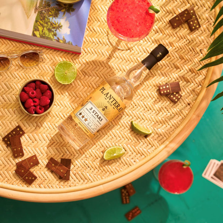 Bottle of Planteray 3 Stars rum on a woven surface with limes, raspberries, and dominoes.