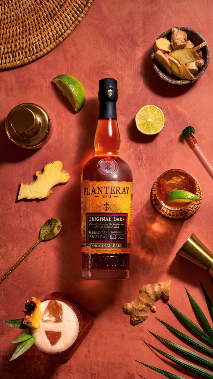 Bottle of Planteray Original Dark Rum on a red surface with garnishes and a woven mat.