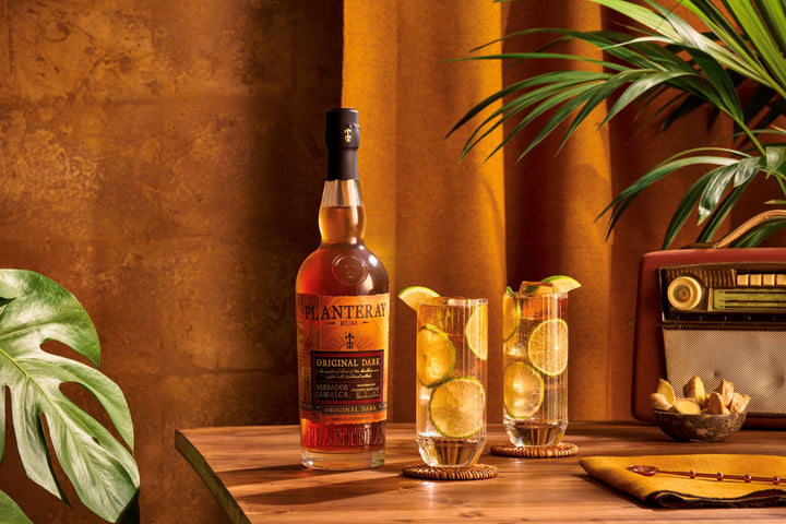Bottle of Planteray Original Dark Rum on a wooden table with cocktails and decorative elements.