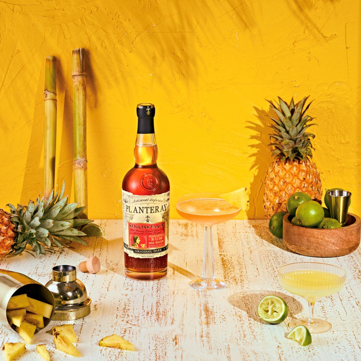 Bottle of Planteray Rum Manhattan cocktail with pineapple, lime, and a glass on a yellow background