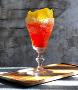 Cocktail with a red and yellow layer in a glass on a wooden tray against a textured wall.