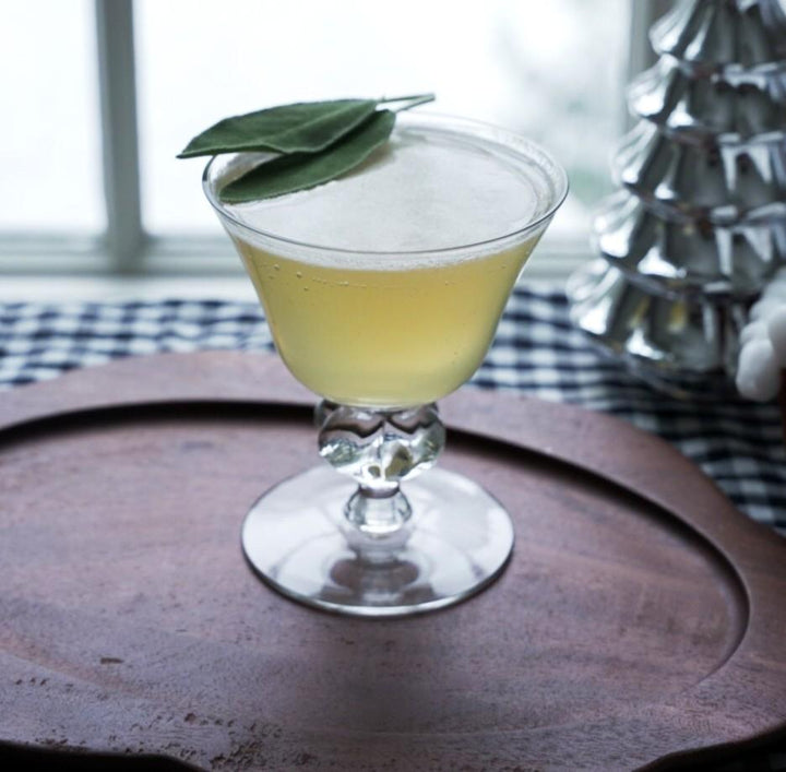 Cocktail in a glass with a leaf garnish on a wooden tray.
