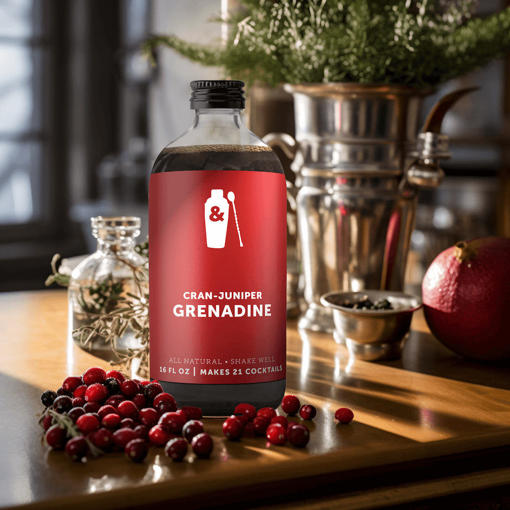 A bottle of Shaker & Spoon Cran-Juniper Grenadine in a kitchen setting, with fresh cranberries, pomegranate, and various spices and flavors arranged artfully around it.