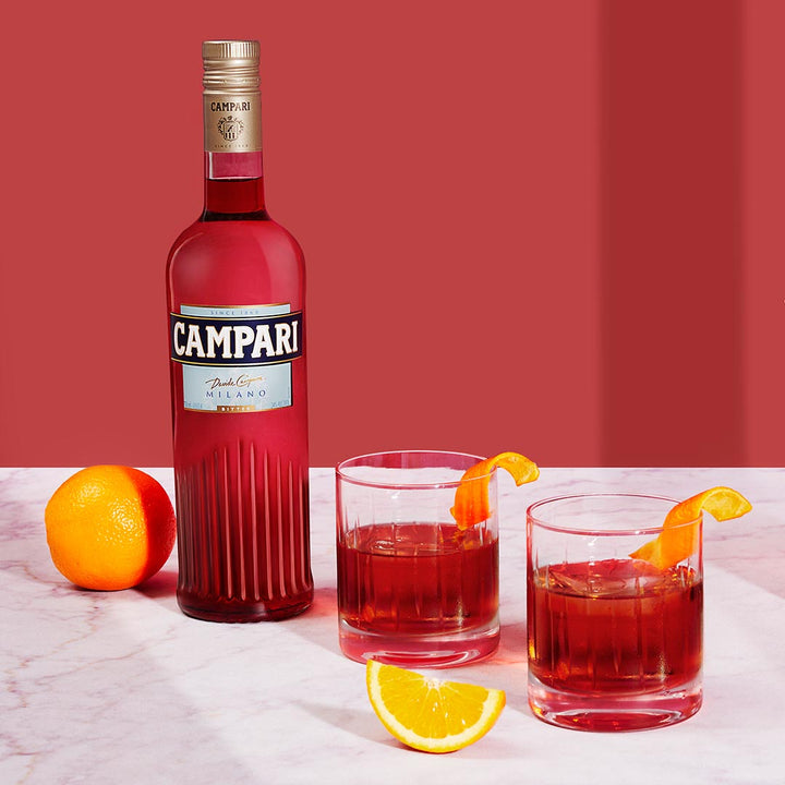 Campari bottle with two glasses of red cocktail and orange slices on a pink background
