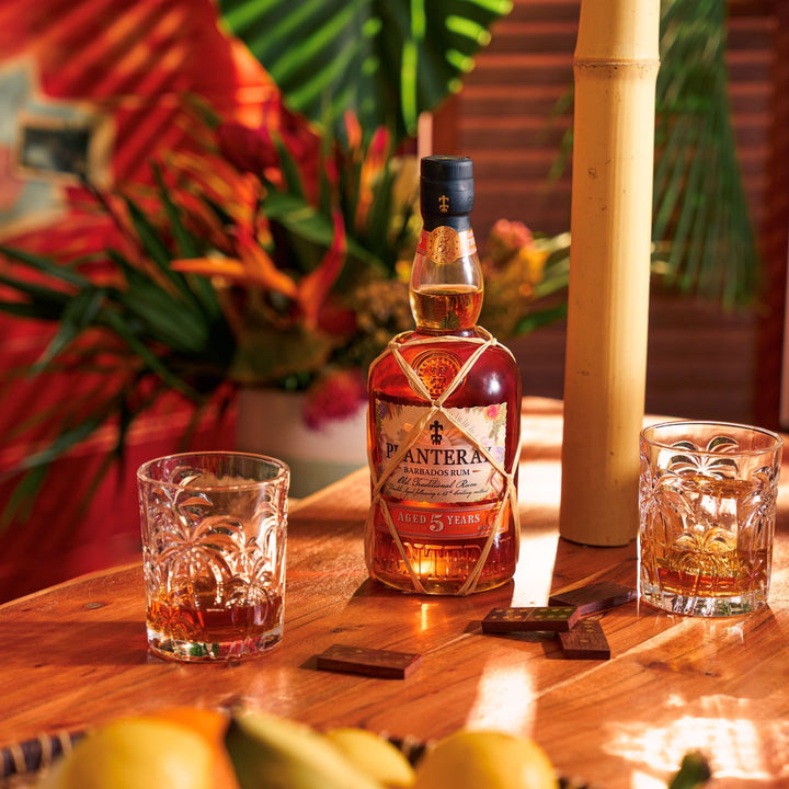 Bottle of Planteray 5 Year Rum with two glasses on a wooden table, surrounded by plants.