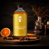 A bottle of Shaker & Spoon Spiced Old Fashioned Syrup in a darkly lit living room setting, with orange slices, cinnamon sticks, and various spices and flavors arranged artfully around it.