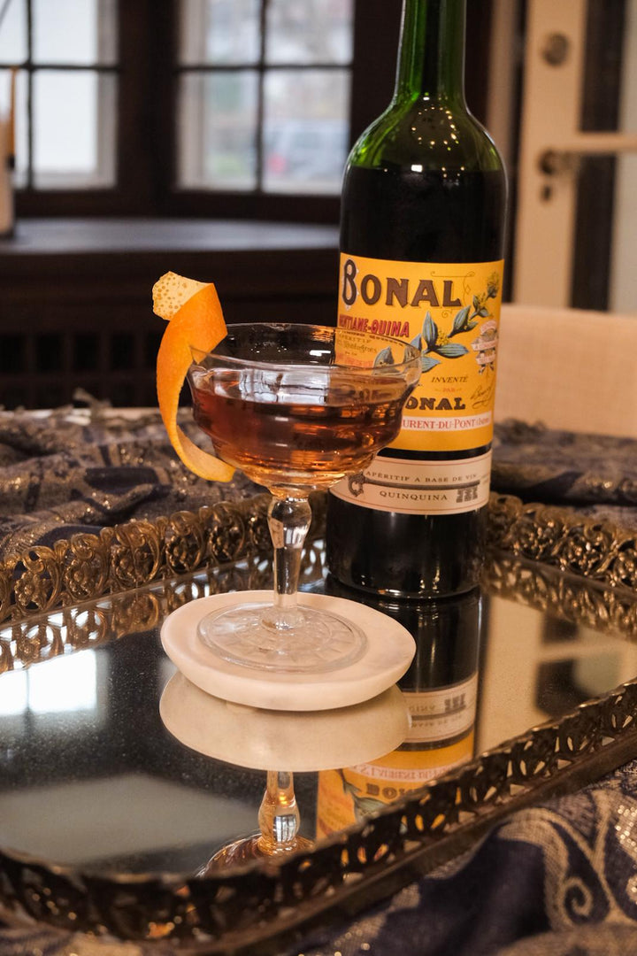 Bottle of Bonal with a cocktail garnished with an orange peel on a reflective surface.