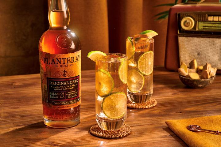 Bottle of Planteray Original Dark Rum with two cocktails garnished with lime on a wooden table.