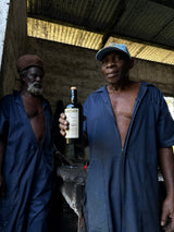 Two men in blue coveralls standing under a corrugated metal roof, one holding a bottle of Jim Beam bourbon.