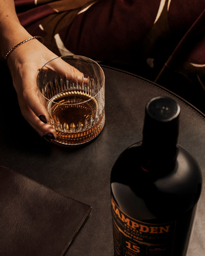 Hand holding a glass of whiskey next to a Hampden bottle on a dark surface