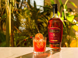 El Dorado 8 Year Old Rum bottle with a cocktail garnished with an orange slice on a table with a blurred green plant background.