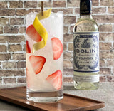 Glass of cocktail with strawberries and lemon peel next to a bottle of Dolin Vermouth on a wooden tray.
