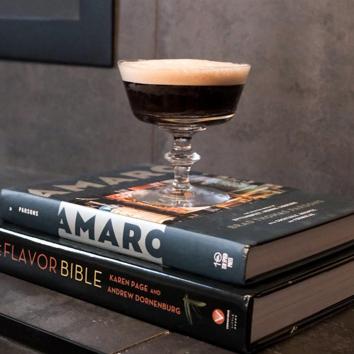 Cocktail glass with a layered drink on top of two books titled 'Amaro Flavor Bible' and 'Parragon'.