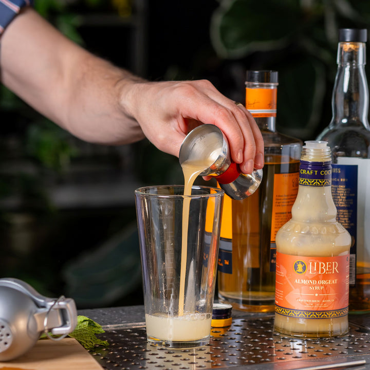 Person pouring a liquid from a spoon into a glass with bottles in the background