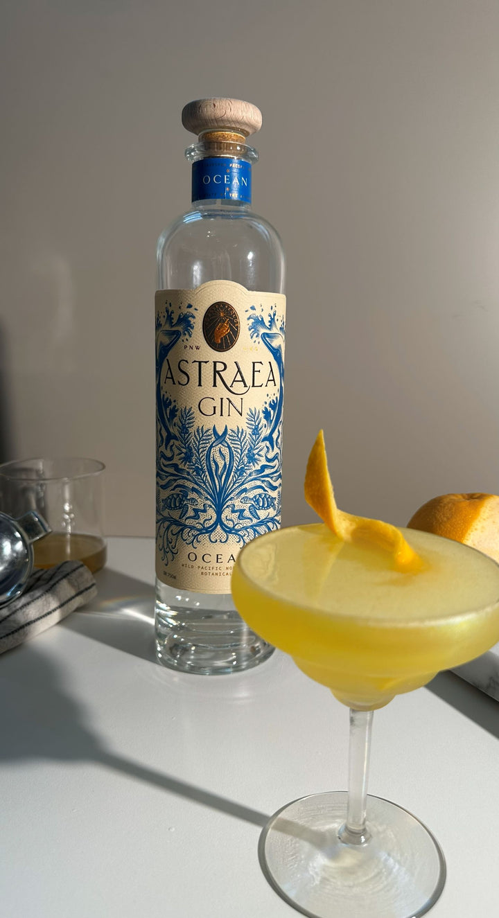 Astraea Gin bottle with a cocktail garnished with a lemon twist on a white surface.