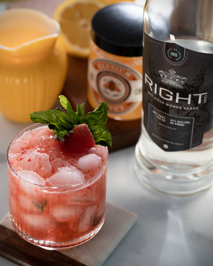 Strawberry cocktail with ice and garnish next to a bottle of gin on a marble surface.