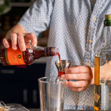 Person pouring a red liquid from a bottle into a cocktail shaker with a blurred background