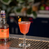 Cocktail in a glass with a bottle of Liber Blood Orange Syrup on a bar counter.