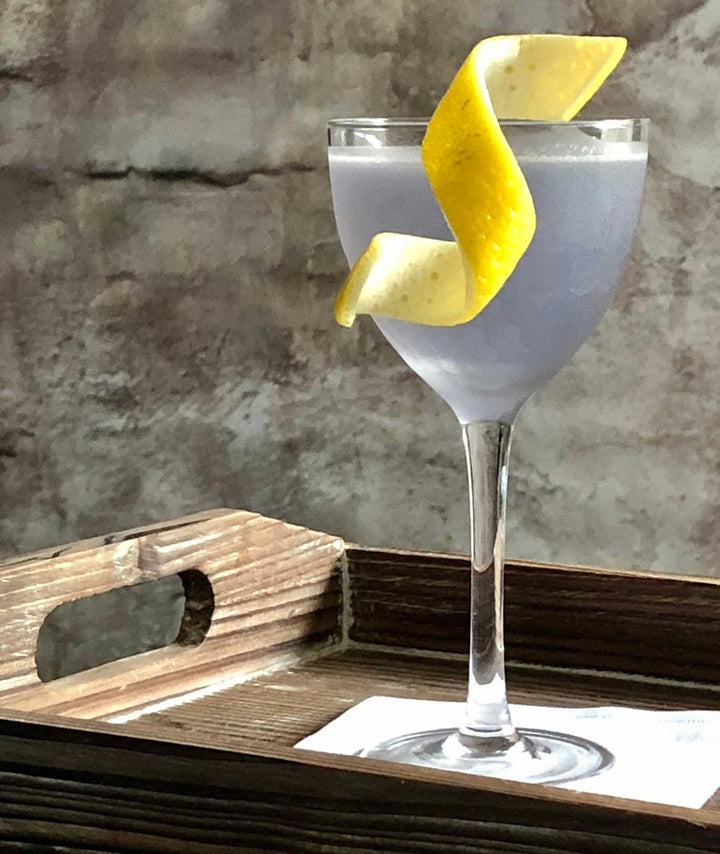 Cocktail with lemon twist in a glass on a wooden tray against a textured wall.